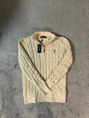 Beige kabelstickad tröja Polo Ralph Lauren - Kabelstickad beige tröja från Polo Ralph Lauren med halv dragkedja och klassisk marinblå logga broderad på bröstet. Tröjan har ribbade muddar och är tillverkad i mjukt material som passar perfekt för kyligare dagar.