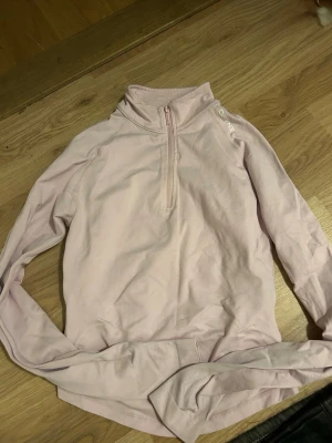Dfyne Impact Half Zippy - Ljusrosa långärmad träningströja från DFYNE med halv dragkedja framtill och hög krage. Tillverkad i ett mjukt och stretchigt material som passar perfekt för träning eller löpning. Diskret logga på axeln och ribbade muddar vid ärmslut.