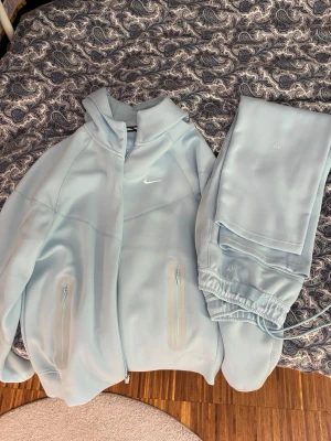 Ljusblå NOCTA Nike hoodie - Säljer en stilren ljusblå hoodie från Nike NOCTA-kollektionen. Hoodien har dragkedja framtill, två fickor med dragkedja och en klassisk huva. Materialet är mjukt och skönt, perfekt för chill dagar. Diskret Nike-logga på bröstet och NOCTA-detaljer.