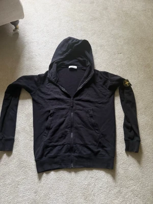 Svart hoodie från Stone Island - Snygg svart hoodie från Stone Island med dragkedja framtill och två fickor. Klassisk huva och ikonisk logotyp på vänster ärm. Tillverkad i mjukt bomullsmaterial som känns skönt mot huden. Perfekt för en avslappnad streetwear-look. Fint skick, pris kan diskuteras