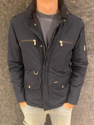 Snoot field jacket - Stilren mörkblå vindjacka från Snoot med flera fickor framtill, dragkedjor i silver och hög krage. Jackan har snygga detaljer som metallknappar, D-ring och en diskret logga på insidan. Perfekt för dig som vill ha en sportig och modern look. Snygg svart quiltad dunjacka med flera fickor framtill, både med knappar och dragkedja. 