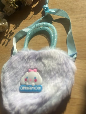Ljuslila fluffig Cinnamoroll handväska - Supersöt handväska i ljuslila fluffigt material med Cinnamoroll-motiv framtill. Väskan har ett hjärtformat utseende, glittriga blå handtag och ett matchande blått axelband. Perfekt för dig som gillar gulliga accessoarer och kawaii-stil.