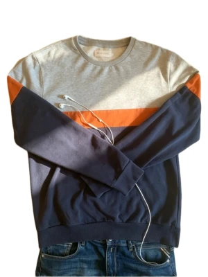 Flerfärgad sweatshirt från Pier One - Snygg sweatshirt från Pier One med färgblock i grått, marinblått och orange. Tröjan har rund halsringning och långa ärmar. Perfekt för en avslappnad och trendig stil. Tillverkad i mjukt bomullsmaterial.