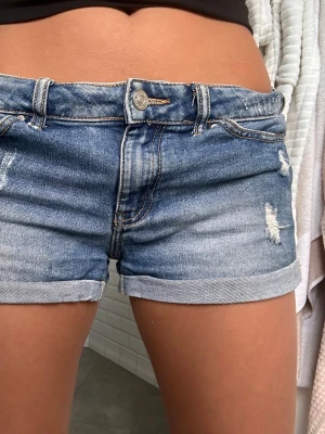 Lågmidjade blå jeansshorts - Säljer ett par lågmidjade blå jeansshorts med slitna detaljer och upprullade benslut. Klassisk femficksmodell med knapp och dragkedja framtill. Perfekta för varma dagar och enkel att matcha med olika toppar.