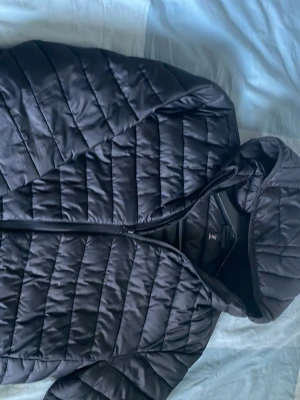Svart pufferjacka med huva, M - Säljer en svart pufferjacka med huva och dragkedja. Jackan har ett quiltat mönster med horisontella sömmar och är tillverkad i ett glansigt material som ger en modern look. Perfekt för kalla vinterdagar och riktigt skön att bära.