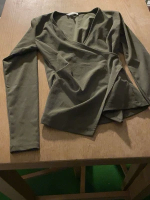Olivgrön omlottopp med lång ärm - Snygg olivgrön omlottopp med lång ärm och v-ringad hals. Toppen har en figurnära passform och är tillverkad i mjukt bomullsmaterial. Perfekt att styla med jeans eller kjol för en trendig look.