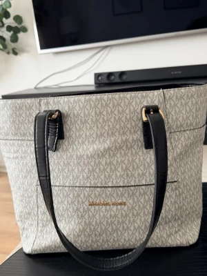 Vit/ grå Michael Kors handväska - Snygg beige handväska från Michael Kors med klassiskt MK-mönster över hela väskan. Väskan har svarta handtag i läderimitation och guldfärgade detaljer. Perfekt storlek för att få plats med allt du behöver och stängs med dragkedja upptill.