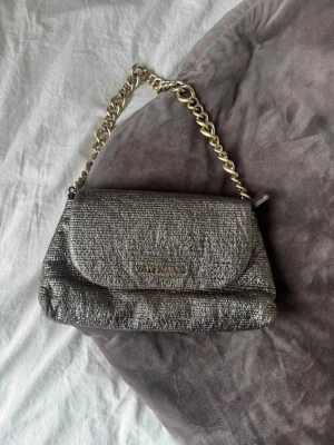 Metallic axelväska med kedja - Snygg metallic valentino axelväska med stor guldfärgad kedja som handtag. Väskan har en strukturerad yta och ett lock med magnetknäppning samt innerfack med dragkedja. Använd 1 gång så den är i helt nyskick.