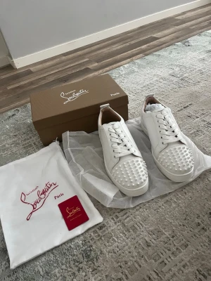 Vita sneakers med nitar från Louboutin - Säljer ett par vita sneakers från Christian Louboutin med coola nitar på tån. Skorna har klassisk låg profil, vita skosnören och är tillverkade i slätt läder. Kommer med originalkartong och dustbag. Perfekta för dig som vill sticka ut med en lyxig touch.
