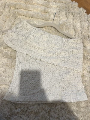 Vit spets oneshoulder topp - Säljer en vit oneshoulder topp från Pacific Republic i spetsmaterial. Toppen har en volangdetalj över axeln och är ärmlös, vilket ger en trendig och somrig look. Perfekt för dig som vill sticka ut lite extra med en unik topp.