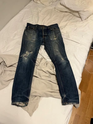 Mörkblå slitna jeans med raka ben - Säljer ett par mörkblå jeans med raka ben och slitningar på både fram- och baksida. Jeansen har trasiga detaljer vid knä och benslut samt klassiska fem fickor. Perfekta för en avslappnad och trendig look.