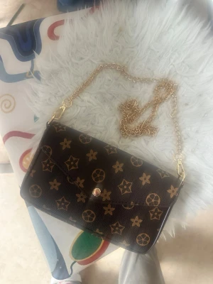 Brun handväska med guldkedja - Snygg brun handväska med guldfärgade stjärn- och cirkelmönster samt guldfärgad kedjerem. Väskan har kuvertform och stängs med en knapp framtill. Perfekt accessoar för att lyfta din outfit.
