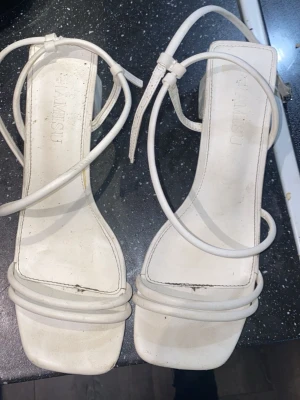 Vita sandaler med remmar från New Yorker - Säljer ett par vita sandaler från New Yorker med tunna remmar och fyrkantig tå. Sandalerna har en stilren design med dubbla remmar framtill och justerbar ankelrem. Tillverkade i syntetmaterial och passar perfekt till sommarens outfits.