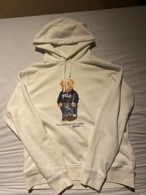 Vit hoodie med Polo Bear från Ralph Lauren - Säljer en vit hoodie från Ralph Lauren med ikoniska Polo Bear-trycket på bröstet. Med de slitna trycket