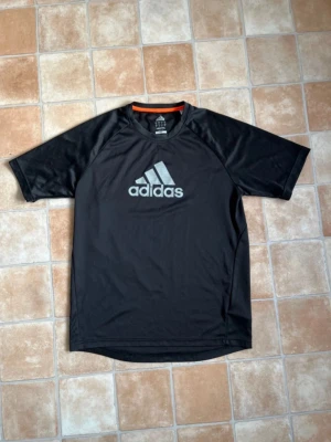 Svart tränings-tshirt från Adidas - Snygg svart tränings-tshirt från Adidas med stor logga på bröstet. Tillverkad i ett lätt och snabbtorkande material som passar perfekt till gymmet eller löprundan. T-shirten har korta ärmar och rund halsringning med en orange detalj på insidan.