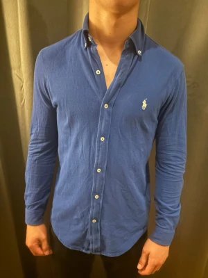 Ralph Lauren skjorta - Snygg blå skjorta från Ralph Lauren med klassisk krage och vita knappar framtill. Skjortan har lång ärm och den ikoniska loggan broderad på bröstet. Tillverkad i mjuk bomull för en bekväm känsla och passar perfekt till många olika stilar.