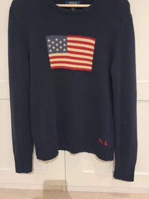 Mörkblå stickad tröja Polo Ralph Lauren - Mörkblå stickad tröja från Polo Ralph Lauren med amerikansk flagga i rött, vitt och blått på bröstet. Tröjan har rund halsringning, ribbstickade muddar och RL-broderi nedtill. Tillverkad i bomull. Köpt på kidsbrandstore så storlek XL i juniorstorlek. Knappt använd 