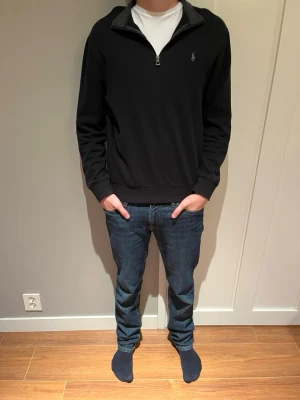 Svart halvzip fleece från Polo Ralph Lauren - Klassisk svart tröja med halv dragkedja från Polo Ralph Lauren. Tröjan har en diskret logga broderad på bröstet och ribbade muddar vid ärmslut och nederkant. Perfekt till jeans eller chinos för en clean och stilren look.