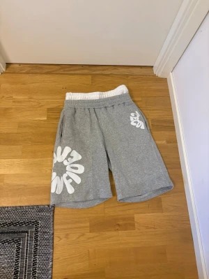 Grå sweatshorts med vit tryck - Säljer ett par grå sweatshorts med bred resårmidja och vita grafiska tryck på ena benet och bak. Shortsen har sidofickor och är tillverkade i mjuk bomullsmix för maximal komfort. Perfekta för chill dagar eller streetwear-stil.