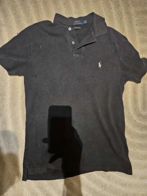 Svart pikétröja från Polo Ralph Lauren - Klassisk svart pikétröja från Polo Ralph Lauren med korta ärmar och vit broderad logga på bröstet. Tröjan har krage och knäppning med två knappar framtill. Perfekt för en stilren och avslappnad look.