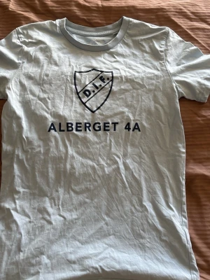 Vit t-shirt med DIF-tryck - Vit t-shirt med blått DIF-märke och texten 'ALBERGET 4A' på bröstet. Klassisk rund halsringning och korta ärmar. Perfekt för dig som vill visa ditt stöd för Djurgården på ett stilrent sätt.