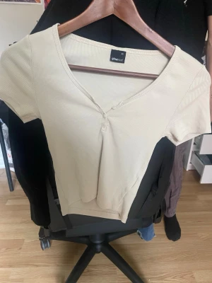 Beige ribbad topp från Gina Tricot - Snygg beige ribbad topp från Gina Tricot med v-ringning och knappar framtill. Kortärmad och i mjukt, stretchigt material som sitter skönt på kroppen. Perfekt att matcha med jeans eller kjol för en enkel och clean look.