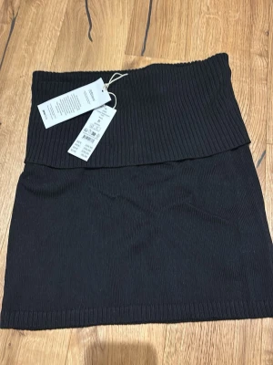 Svart ribbad stickad kjol - Svart stickad kjol med ribbad struktur och bred, nedvikt midja. Kjolen är tillverkad i ett mjukt material och har en enkel, rak passform. Perfekt för att skapa en stilren look med en trendig touch. Kan även användas som en topp. Ny med prislapp, nypris 359kr. 