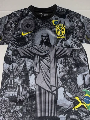 Nike Brasil grafisk fotbollströja - Snygg svart och grå fotbollströja från Nike med Brasil-logga och gult Nike-märke. Framsidan har ett stort grafiskt tryck med Kristusstatyn och brasilianska motiv. Tillverkad i lätt och ventilerande material, perfekt för träning.