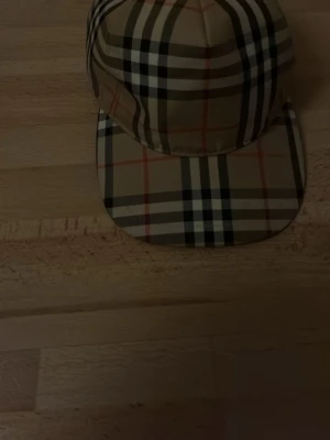Beige burberry  keps med rutigt mönster - Snygg beige burberry  keps med klassiskt rutigt mönster i svart, vitt och rött. Kepsen har böjd skärm och justerbar baksida. Perfekt accessoar för en trendig streetstyle-look.