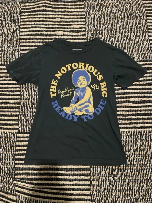Svart The Notorious BIG t-shirt - Cool svart t-shirt med tryck av The Notorious BIG och texten 'Ready To Die' i gult och blått. Klassisk passform med rund hals och korta ärmar. Perfekt för dig som gillar hiphop och streetwear.