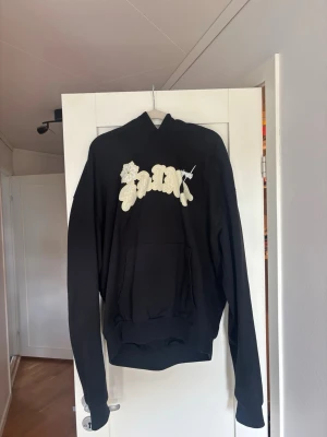 MJGONZALES Hoodie  - Svart MJGONZALES hoodie med stor ficka framtill. På bröstet finns ett stort, krämfärgat tryck med texten 'SAINT' i en grafisk stil och med en liten blomdetalj. Perfekt för dig som gillar streetwear och vill sticka ut med en unik design. Extremt bra kvalité i tyget. 