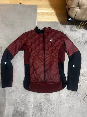 Vinröd sportjacka från Asics - Snygg vinröd och svart sportjacka från Asics med quiltad framsida, hel dragkedja och reflekterande detaljer på ärmarna. Jackan har figurnära passform, hög krage och paneler i stretchigt material för extra rörelsefrihet. Perfekt för löpning eller cykling.