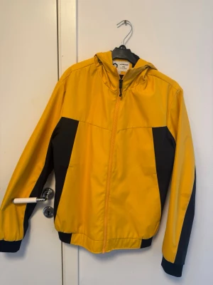 Gul och svart vindjacka Jack & Jones - Snygg gul vindjacka från Jack & Jones med svarta detaljer på sidorna och ärmarna. Jackan har huva, dragkedja framtill och muddar vid ärmslut och midja. Perfekt för blåsiga dagar och enkel att matcha med streetwear.