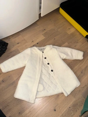 Vit teddyjacka med knappar - Mysig vit teddyjacka med rund hals och svarta knappar framtill. Jackan har quiltat innerfoder och en söt boll-detalj på insidan. Perfekt för kalla dagar när du vill vara både varm och stylish.