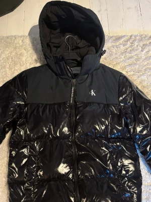 Svart pufferjacka från Calvin Klein - Snygg svart pufferjacka från Calvin Klein med glansig finish och luva. Är mycket bra skick och perfekt som höst/vinterjacka är varm och skön. Storlek M 