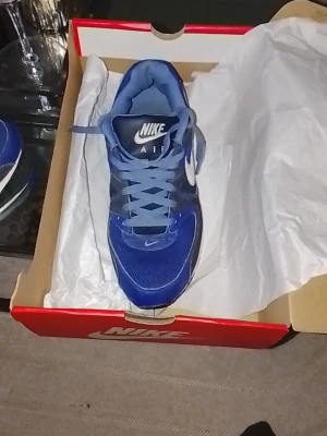 Blå Nike Air sneakers - Snygga blå Nike Air sneakers med vit sula och vit swoosh på sidan. Skorna har meshdetaljer för bra ventilation och klassisk snörning. Den dämpade sulan ger extra komfort och den sportiga designen gör dem perfekta för en aktiv livsstil.