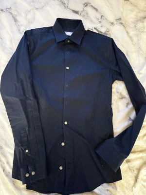 Mörkblå skjorta Tiger of Sweden - Snygg mörkblå skjorta från Tiger of Sweden i extra slim fit. Skjortan har klassisk krage, långa ärmar och vita knappar. Tillverkad i ett tunt och stilrent tyg som passar perfekt till en dressad look.