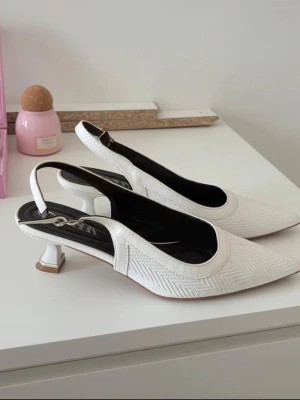 Vita slingback-pumps med låg klack - Säljer ett par vita pumps med elegant slingback-rem och spetsig tå. Skorna har låg, fyrkantig klack och ett diskret fiskbensmönster i materialet. Perfekta för dig som gillar stilrena och trendiga skor med en twist.