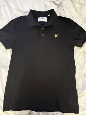 Svart pikétröja Lyle & Scott - Svart pikétröja från Lyle & Scott med klassisk krage, två knappar och gul logga på bröstet. Tillverkad i ekologisk bomull för en skön känsla och stilren look. Passar perfekt till både jeans och shorts.