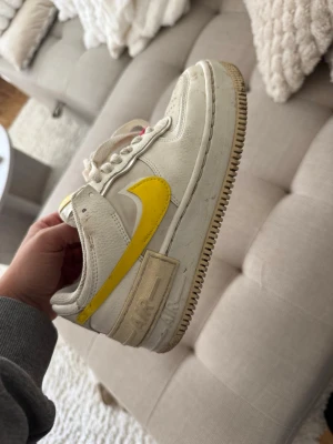 Vita och gula Nike Air Force 1 sneakers - Säljer ett par vita Nike Air Force 1 sneakers med gula detaljer och swoosh. Skorna har klassisk låg siluett, perforerad tå och vita snören med rosa detaljer. Tillverkade i läder och textil för en cool och sportig look.