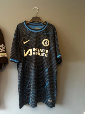 Chelsea FC tredjeställ Gallagher #23 - Säljer en Chelsea FC bortatröja från Nike med Gallagher och nummer 23 på ryggen. Tröjan är svart med blått rutmönster, har klubbmärke och sponsortryck på bröstet samt rund hals och korta ärmar. Perfekt för dig som älskar fotboll och vill stötta Chelsea. Använt ca 2 gånger så inprincip som ny. Säljes då den inte kom till så mycket användning utöver de två tillfällena. 