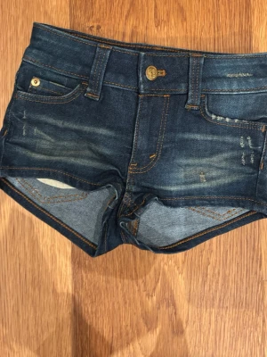 Mörkblå jeansshorts med tryck - Snygga mörkblå jeansshorts med slitningar och kontrastsömmar. Femficksmodell med knapp och dragkedja framtill. På baksidan finns ett grönt tryck med text och döskalle. Materialet är klassisk denim.