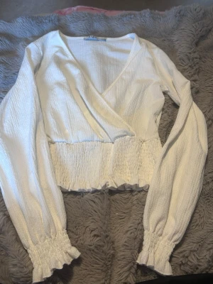 Vit omlottopp från NA-KD - Snygg vit omlottopp från NA-KD med långärmad design och v-ringning. Toppen har smockad midja och ärmslut samt volangdetaljer längst ner. Perfekt för en trendig och fräsch look.
