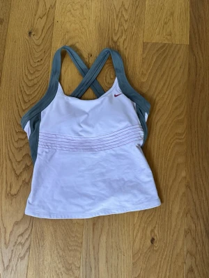 Nike vitt sportlinne med grå detaljer - Snyggt sportlinne från Nike i vitt med grå axelband och sidor. Linnet har korsade axelband i ryggen och tunna röda linjer framtill samt en liten röd Nike-logga. Perfekt för träning och sportaktiviteter.passar m i storleken.