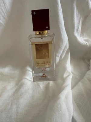 Ana Abiyedh Rouge parfym - Ana Abiyedh Rouge är en elegant parfym i en rektangulär glasflaska med gulddetaljer och en vinröd fyrkantig kork. Flaskan har en lyxig känsla med guldfärgad etikett och arabiska inskriptioner. Tillverkad i Förenade Arabemiraten, Eau de Parfum.