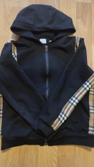 Burberry Hoodie - Snygg svart hoodie med dragkedja och huva. Ärmarna har klassiska beige, röda och svarta rutiga paneler längs sidorna som ger en unik look. Tillverkad i mjukt sweatshirtmaterial. Nytt pris 3000 MITT PRIS 1000kr ENDAST‼️🔥 tags kommer med. OBS KVITTO KOMMER ICKE MED.