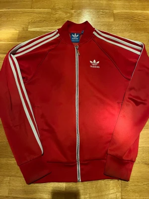 Röd Adidas Originals track jacket - Snygg röd Adidas Originals track jacket i storlek XS med vita ränder längs ärmarna och dragkedja framtill. Klassisk retrostil med Adidas logga broderad på bröstet. 