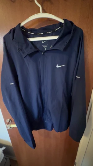 Mörkblå Nike Miller jacka  - Snygg mörkblå vindjacka från Nike med huva och dragkedja framtill. Jackan har långa ärmar och reflekterande detaljer på bröstet och ärmarna. Perfekt för löpning eller vardagsbruk. Lätt och smidig design.