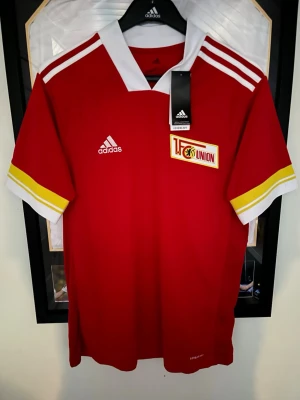 Röd 1. FC Union matchtröja Adidas - Snygg röd matchtröja från Adidas för 1. FC Union Berlin. Tröjan har vita och gula detaljer på ärmarna, vit krage och klubbmärke på bröstet. Tillverkad i lätt och ventilerande material, perfekt för fotboll eller träning.