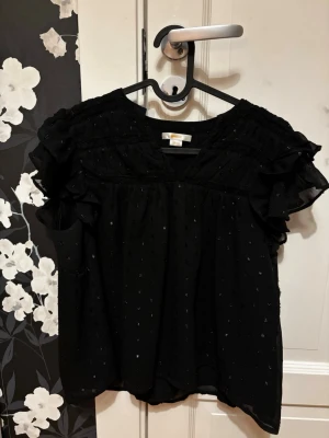 Svart blus med volangärmar - Snygg svart blus i tunn, luftig chiffong med små diskreta prickar och volangdetaljer på ärmarna. Blusen har v-ringning och korta, rynkade ärmar som ger en romantisk vibe. Perfekt att styla med jeans eller kjol för en trendig look.